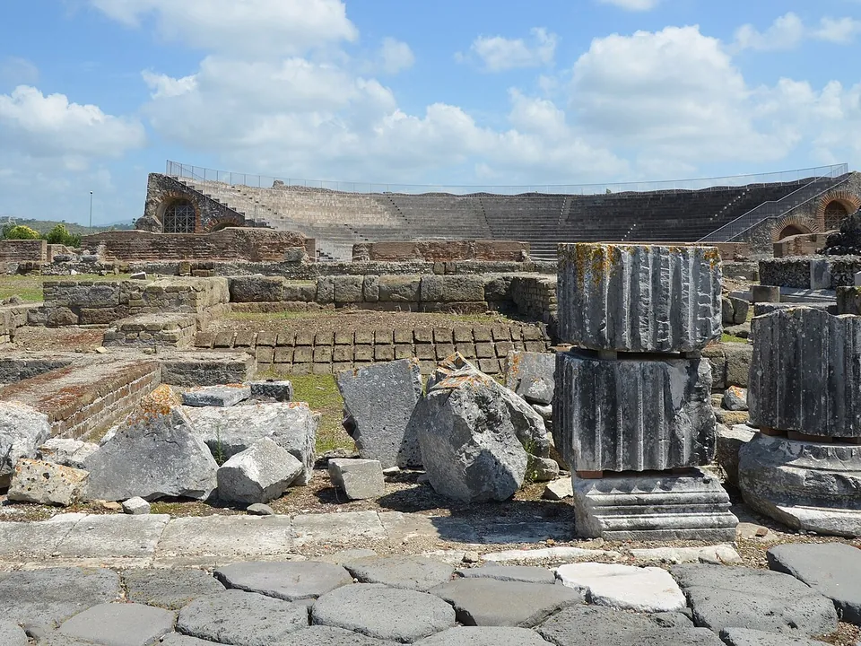 Large tumbled stone blocks and stacked fluted column drums beside a low excavated area, with the curved tiers of the theatre on the skyline.