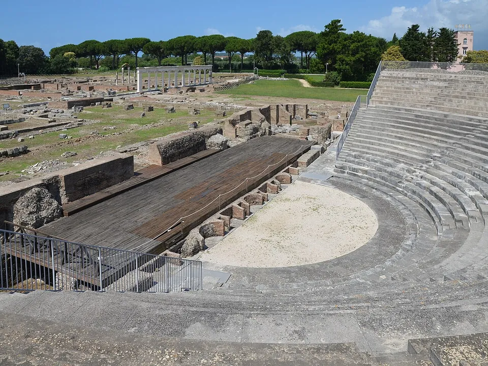 Looking down the curved stone tiers of the theatre to its semicircular orchestra and a wooden stage platform, with excavated walls, a row of columns and pines beyond.