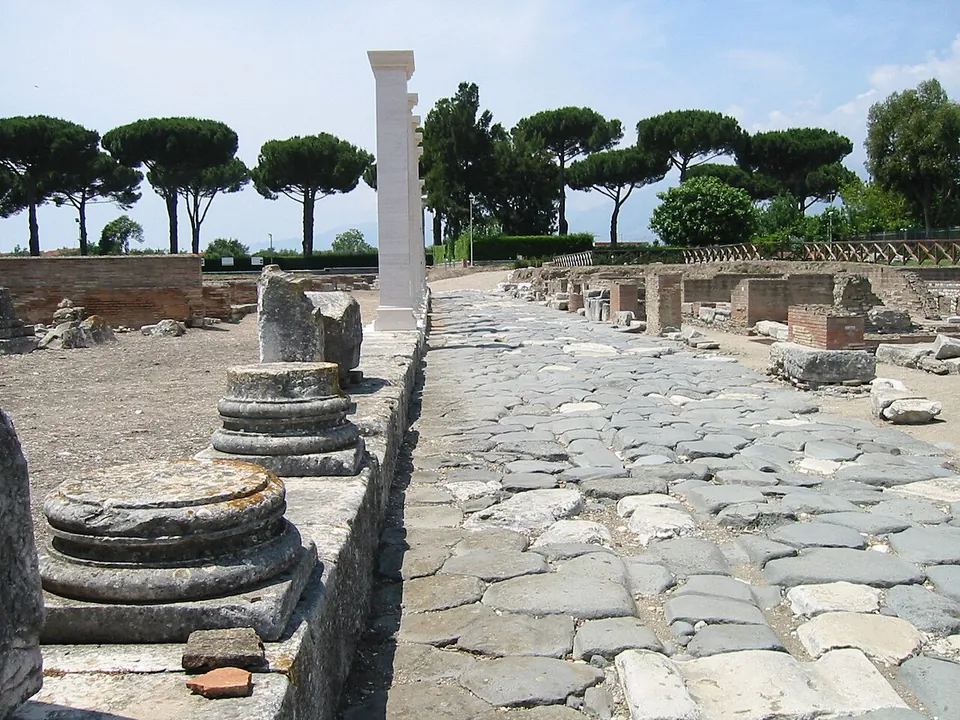 A road paved with large, irregular grey stone blocks running straight between excavated walls, with round column bases on a raised kerb to the left, one tall white column, and umbrella pines in the distance.