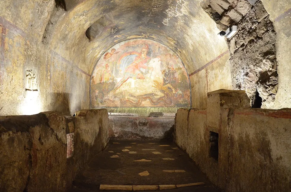 A long, low vaulted underground room with raised benches along both side walls and a large wall painting filling the far end.