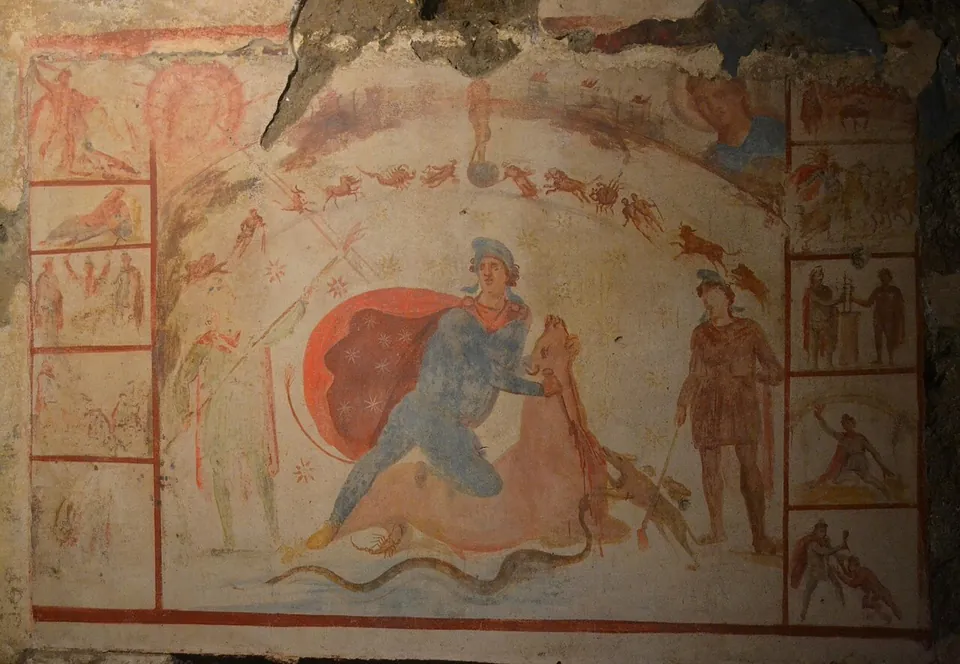 A faded wall painting framed in red lines. In the middle, a figure in a blue tunic and a cap, a red cloak flying behind him, kneels on a bull and holds its head back; a standing figure watches from the right, a dog and a snake reach up at the bull, and an arc of small zodiac figures runs overhead. Narrow columns of small framed scenes run down both sides.