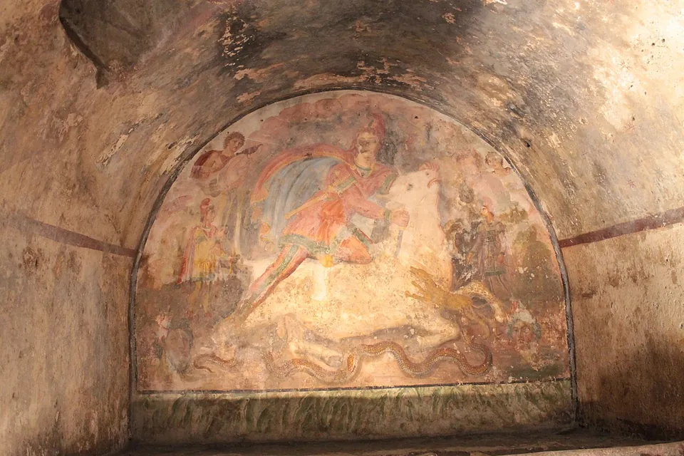 The end wall of a vaulted underground room filled by a faded painting under the arch of the vault - a figure in red with a billowing cloak bears down on a bull, with smaller figures at either side and the upper corners - above a low ledge of masonry.