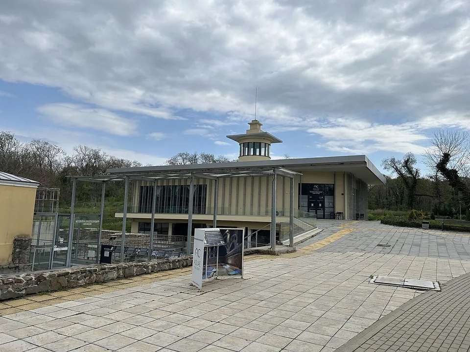 A paved square leading to a modern building with a colonnaded front and a small domed lantern, a canopy over the ruins on the left and an information stand in front.