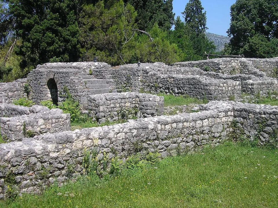 Thick rubble-stone walls about head high, with a small round-arched opening and a flight of stone steps rising along the wall, on a lawn with tall cypresses behind.