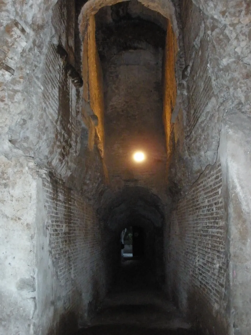 A long, narrow brick-walled passage with a barrel vault, lit by a single lamp, running away from the viewer into darkness.