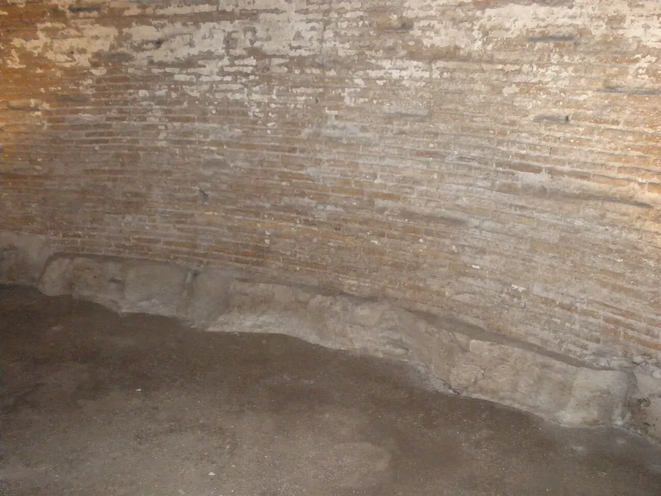 The foot of a curved brick wall, with a band of rough pale stone blocks along its base meeting an earth floor.