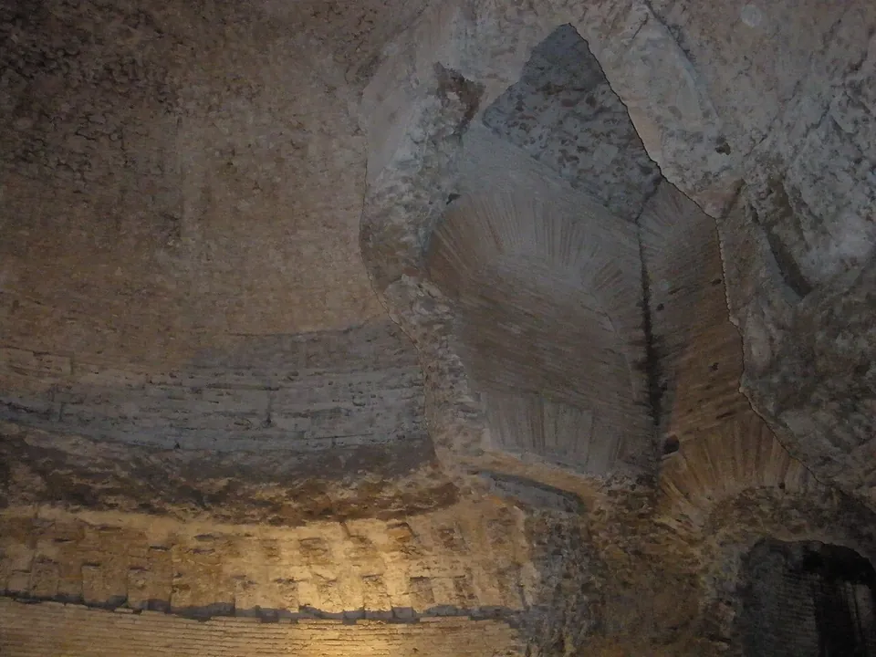 The curved brick wall of a dark circular chamber, with a broken ledge running round it and a pointed opening high in the wall where the masonry has fallen away to show brickwork behind.