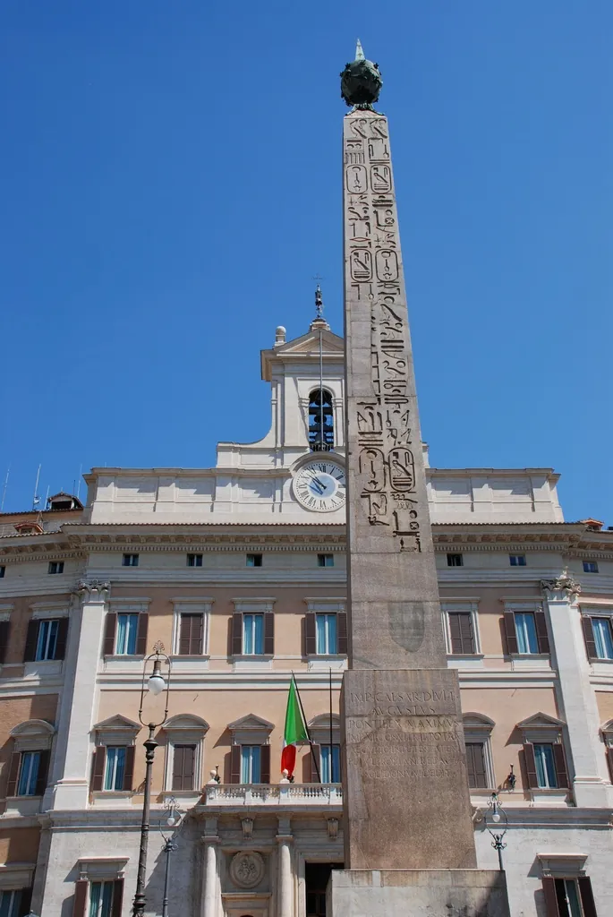 A tall obelisk carved with hieroglyphs in its upper part and topped by a metal globe, standing in front of the pale façade of a palace with a clock and a bell turret, against a clear blue sky.