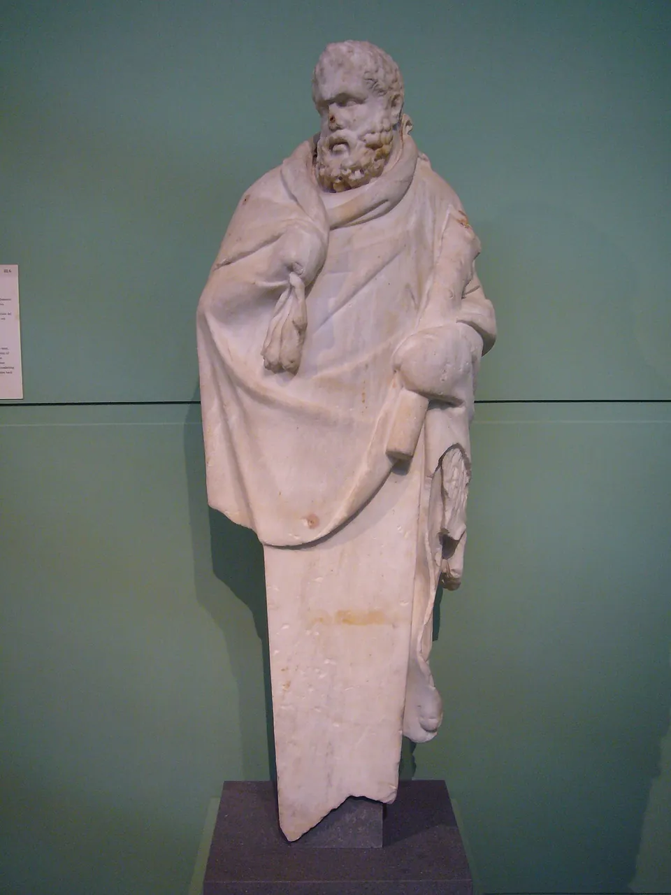 A weathered white marble figure of a bearded man wrapped in a cloak, standing on a dark plinth against a pale green museum wall.