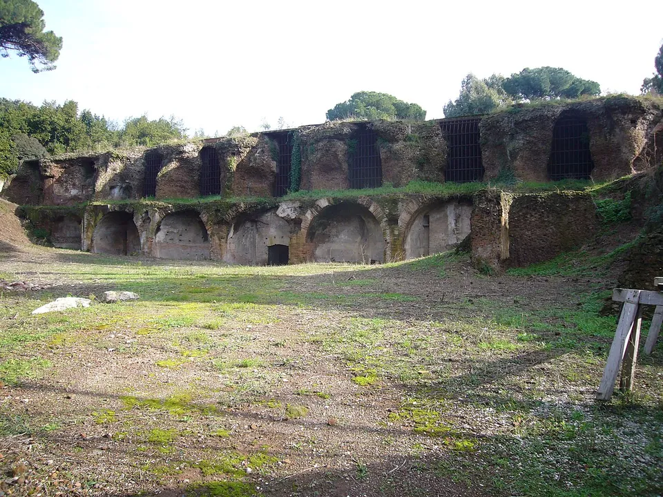 A long ruined brick front with two rows of arched openings, several of the upper ones closed by iron grilles, at the edge of a grassy clearing with trees above.