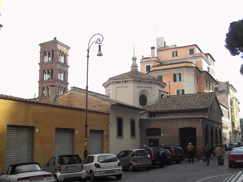 A cobbled street with parked cars below a tall brick bell tower with arched openings on each storey, a small domed building, and on the right a low brick building with round-arched openings along its side.