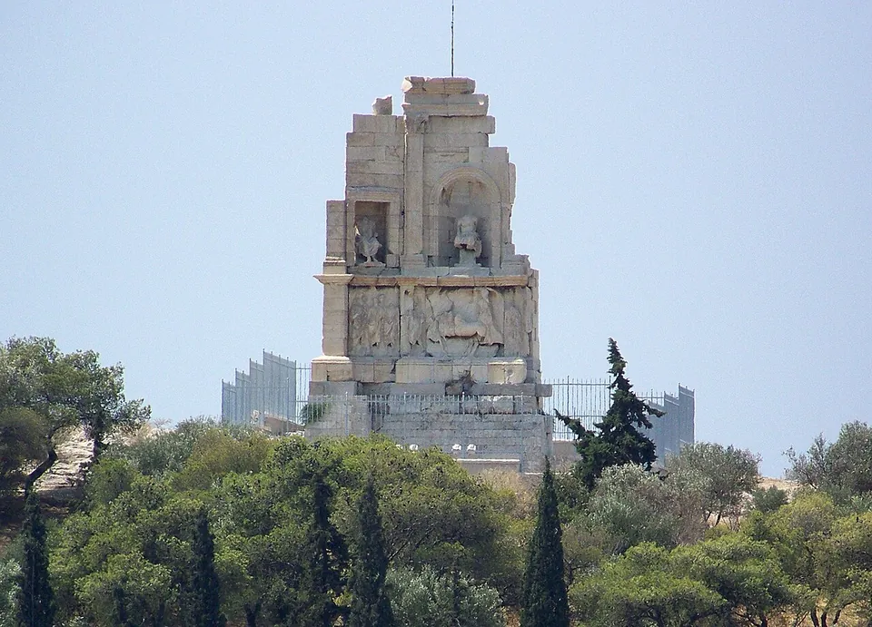 A tall marble monument standing above pines and cypresses, its front holding a seated headless statue in a central niche and another in a niche to the left, with a long carved relief of figures below them, inside a metal fence.