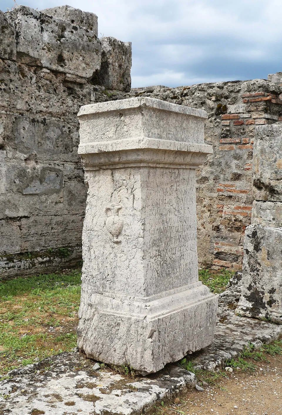 A tall stone pedestal with a moulded top and base, carved on one face with a jug in relief and on another with lines of worn lettering, standing against a wall of stone and brick.