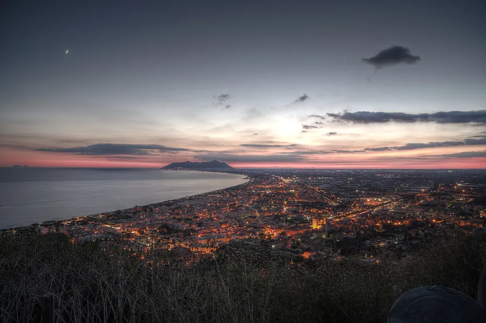 An evening view from high above over the lights of a town spreading along a curving shore, with a mountain on the horizon under a pink and grey sky.