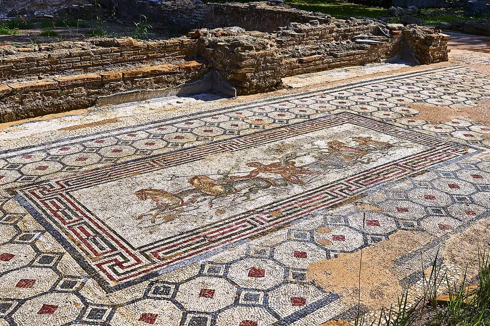 A floor mosaic in a red and black border showing a sea creature riding among horses with fish tails, set in a wider floor of black hexagons, with low brick walls behind.