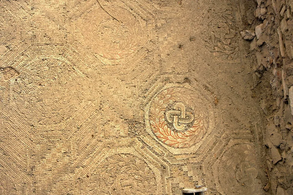 A section of mosaic floor in pale and terracotta tesserae, a pattern of octagons outlined in darker stones, one of them framing a knot motif, with a rough earth edge on the right.