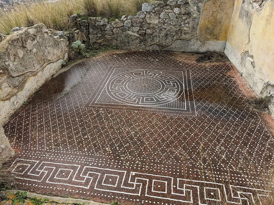 A floor of small dark tesserae dotted with a lattice of white ones, with a square panel framing a circle of interlocking patterns at its centre and a white meander border along the near edge, between low rubble walls.
