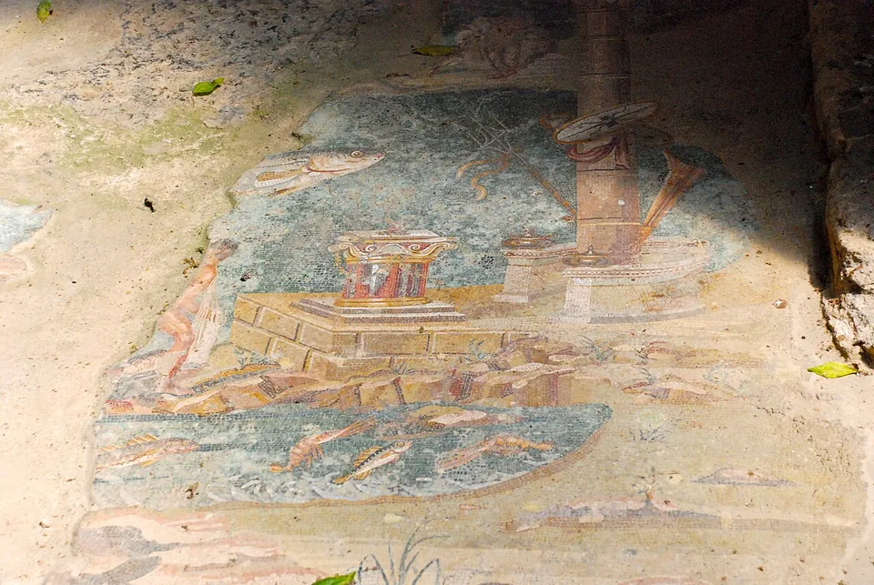 A worn floor mosaic in blues, greens and reds, with fish swimming around a small red altar-like block and a tall column standing on a shore, its surface patched and broken in places.