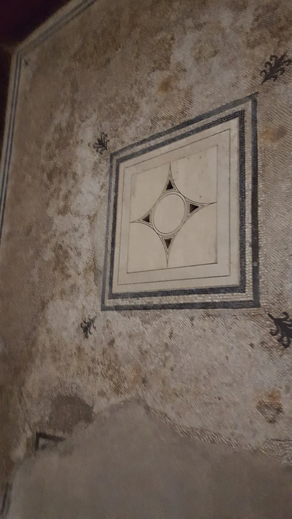 A pale mosaic floor, worn and patchy, with a square panel framed by black and white bands enclosing a white field with a black four-pointed star around a circle.