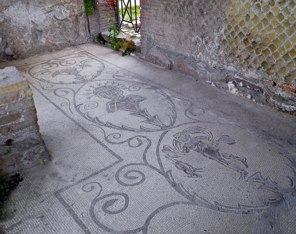 A black-and-white mosaic floor of scrolling tendrils framing figures, laid in a narrow room whose wall shows a net pattern of small square stones.