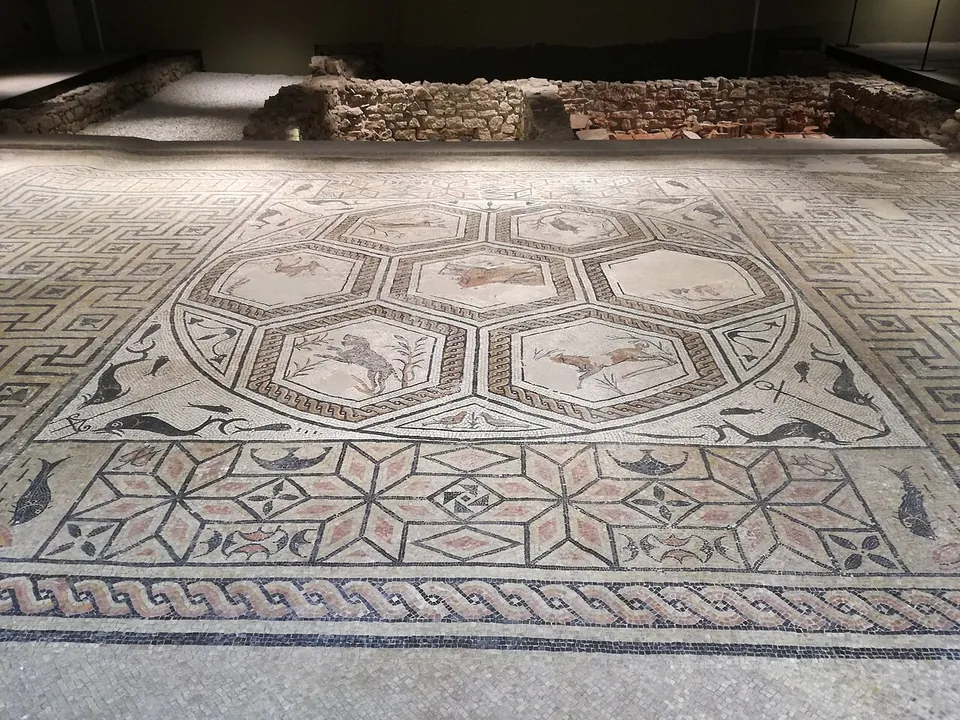 A large mosaic floor under low lighting, its centre a ring of hexagonal panels showing running animals, with dolphins in the corners, a band of eight-pointed stars below and a twisted-rope border, and low stone walls behind it.