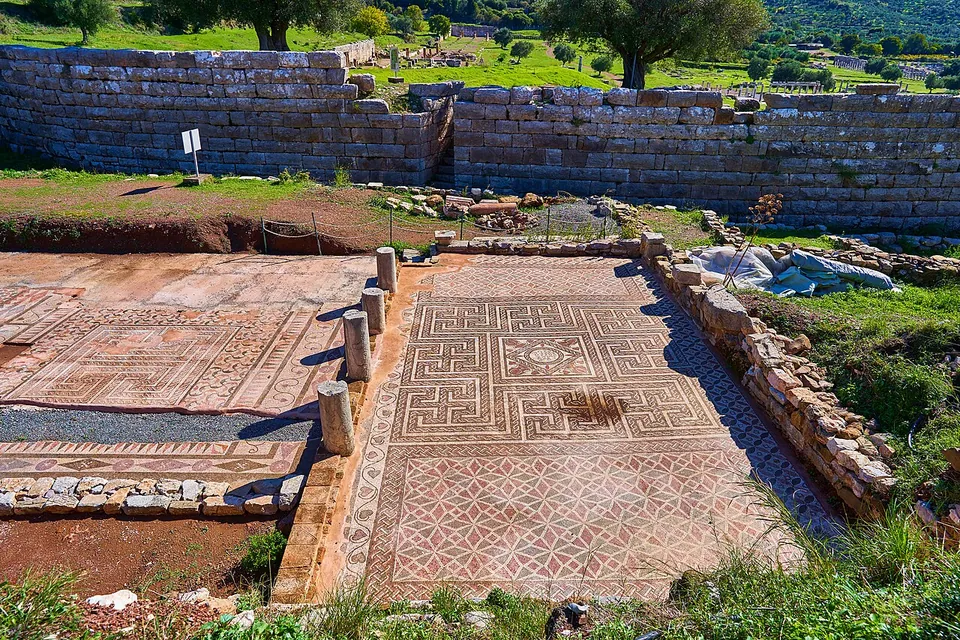 Two large mosaic floors with geometric patterns of squares, meanders and lozenges, separated by a row of short column stumps, below a wall of big squared blocks.