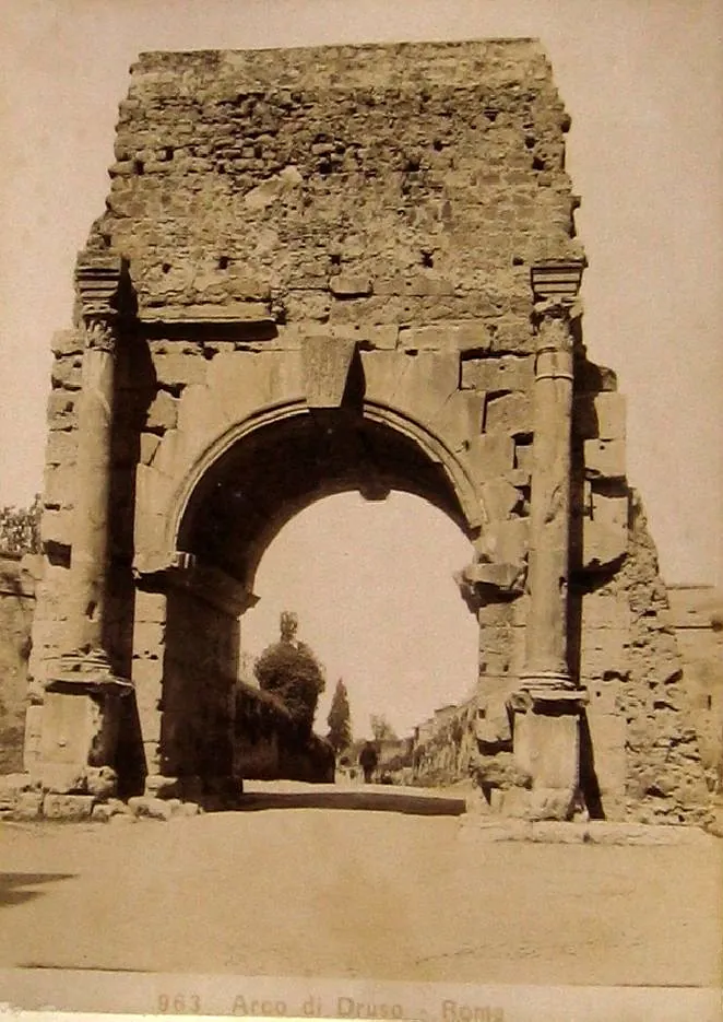An old sepia photograph of a single stone arch framed by two columns, its upper part a mass of rough masonry, with a road passing through it and a domed building in the distance.