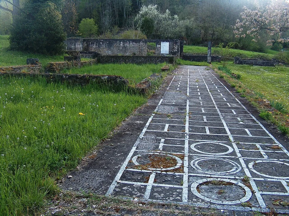 Low conserved stone walls of a villa in a meadow, with a long strip of floor patterned in a white grid with circles running along one side towards a higher wall carrying a sign, and flowering trees behind.