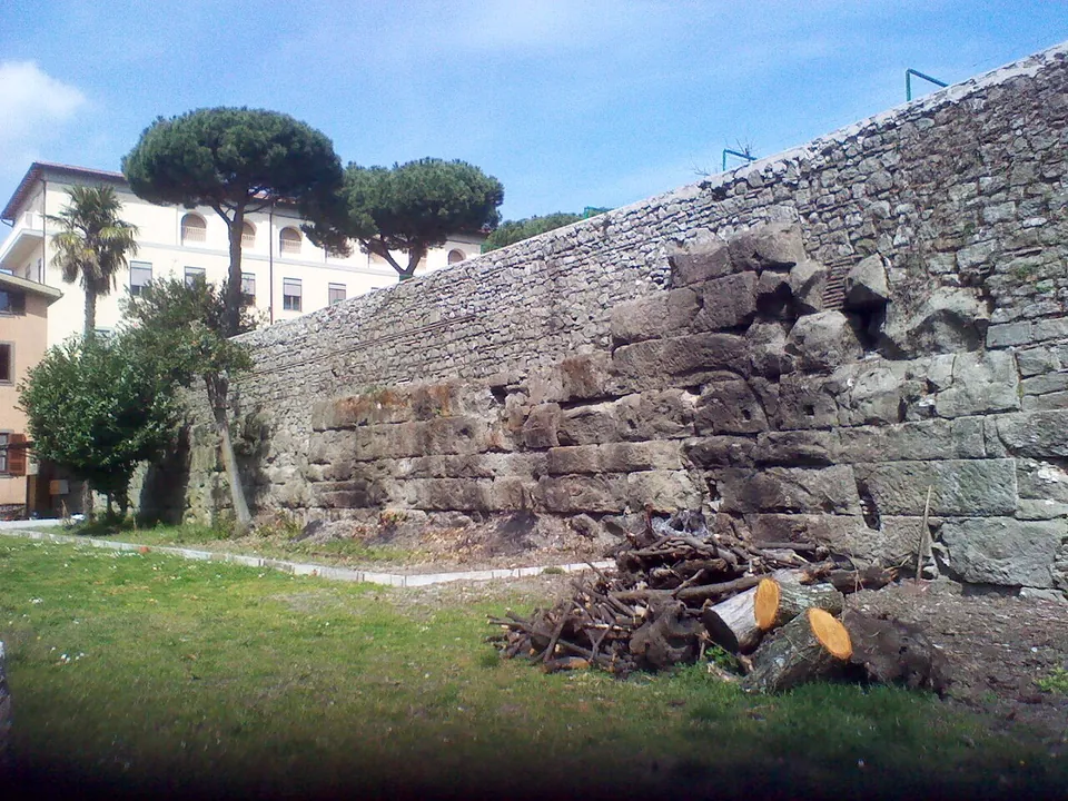 A long, straight wall of squared stone blocks laid in regular courses, standing beside a lawn with umbrella pines and modern buildings behind it.
