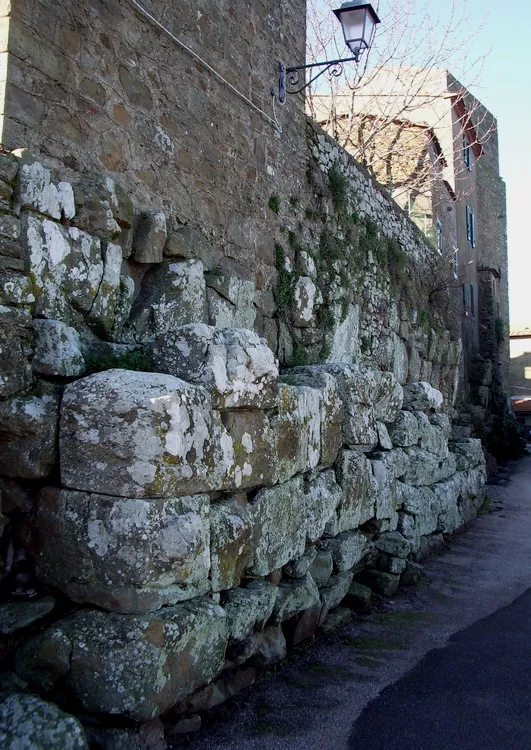 Huge rough-cut limestone blocks, grey and lichen-covered, form the lower part of a wall beside a narrow village street, with later masonry and a street lamp above.