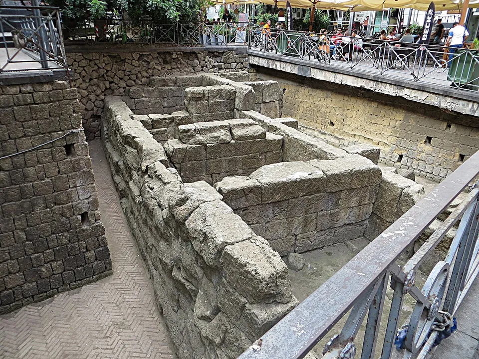 Walls of large squared tuff blocks at the bottom of an open excavation pit, a few metres below a railing with café tables and umbrellas at street level.