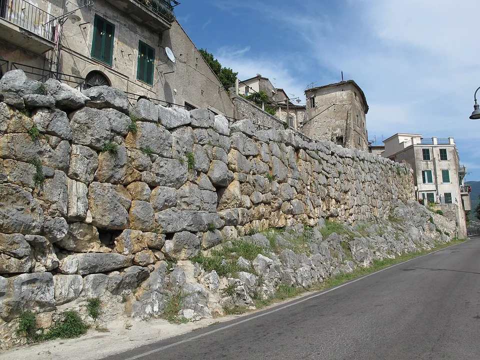 A long wall of large, irregular grey limestone blocks fitted together runs beside an asphalt road, with houses and balconies built on top of it.