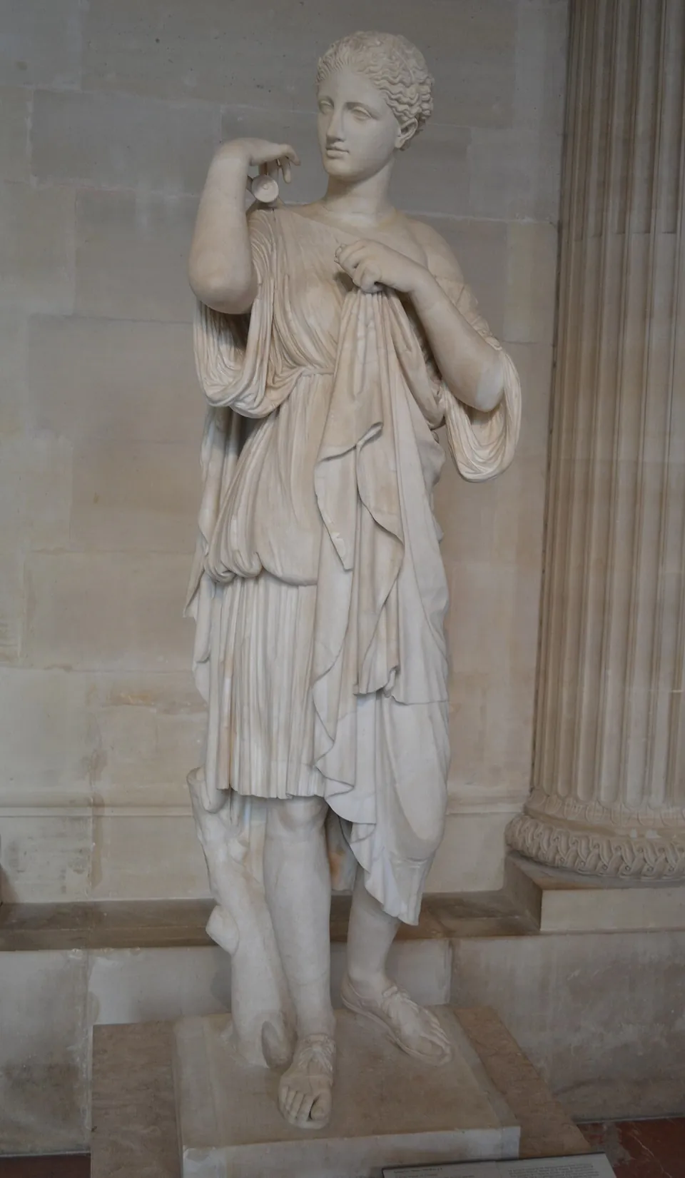 A marble statue of a young woman in a short belted tunic, fastening a cloak at her shoulder, standing in a gallery beside a fluted column.