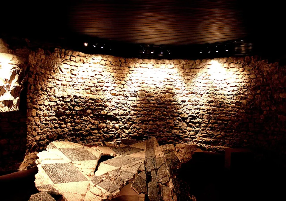 A curved wall of rough stones lit by spotlights in a dark room, with broken slabs of a black-and-white patterned mosaic floor lying in front of it.
