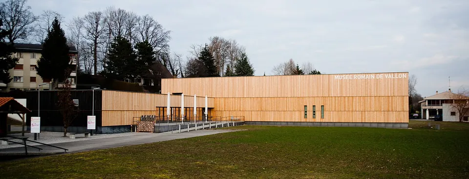 A long, low museum building clad in pale wooden boards, with its name lettered along the top of the façade, beyond a lawn and a paved forecourt.