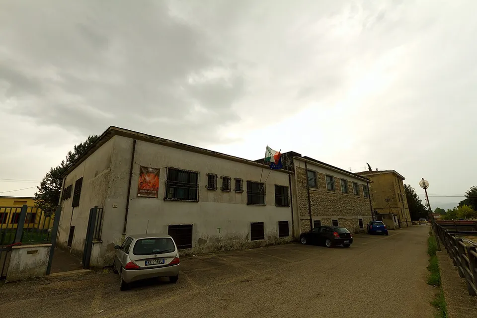 A plain two-storey modern building with a flag over the entrance and cars parked in front, under a grey sky.