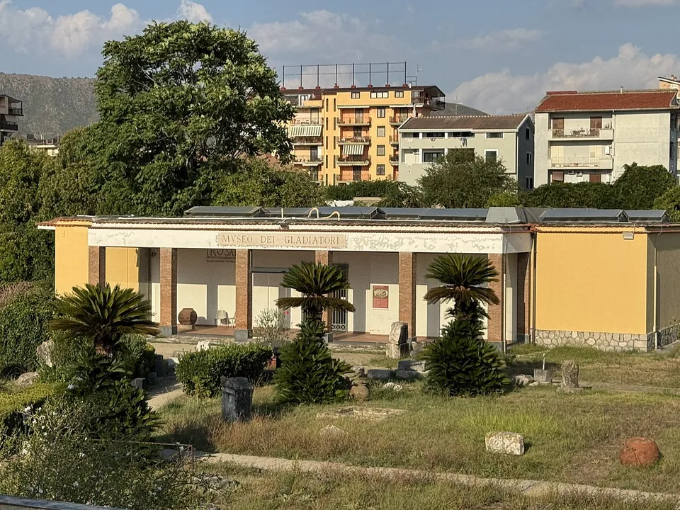 A low yellow building with a columned porch and the words Museo dei Gladiatori over it, in a garden of palms and grass with fragments of stone set along the paths.