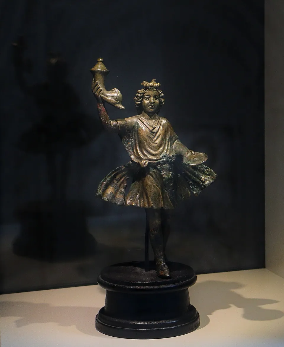 A small bronze statuette of a dancing boy in a belted, flaring tunic, one arm raised high holding a vessel, on a round black base in a dark display case.