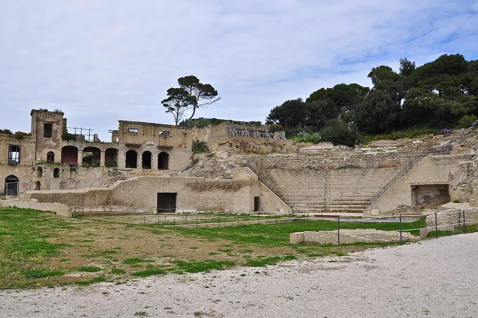 Tiers of stone seating rising in a semicircle against the hillside, below a ruined two-storey building with a row of arched openings and pine trees on the crest, with a lawn and a gravel path in front.