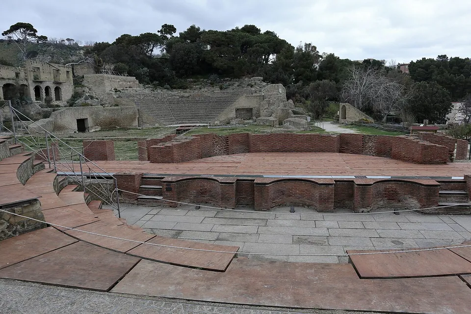 A wide, curved view across a small semicircle of low brick-red seating and a paved stage in the foreground, with a much larger bank of stone seating cut into a wooded slope behind it and ruined buildings above.