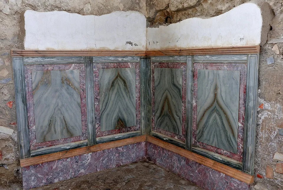 The corner of a room whose lower walls are faced with framed panels of green, wavy-veined decoration above a purple-red base, the plaster above them broken away.