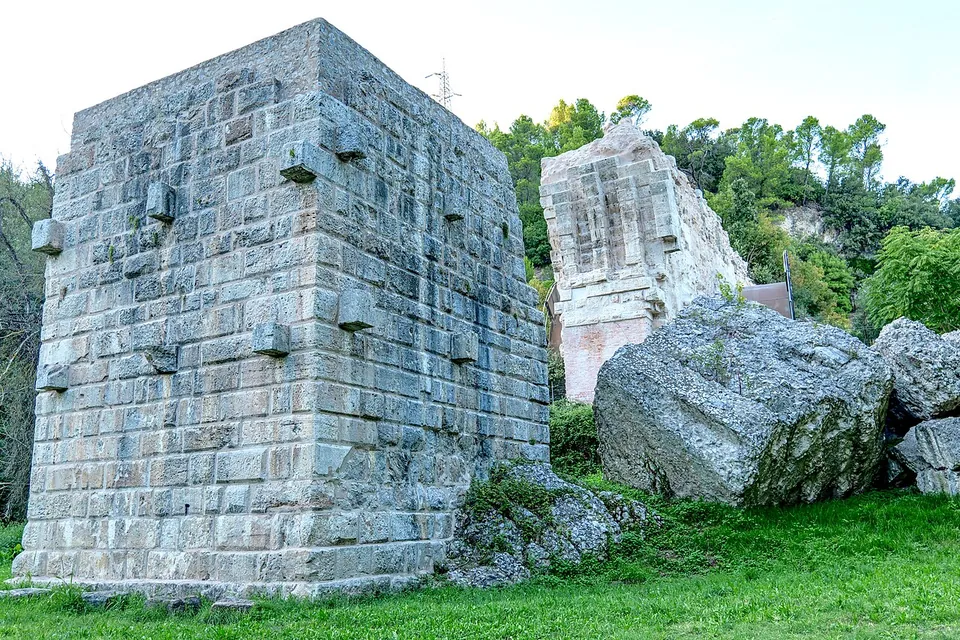 A free-standing block of masonry built of squared pale stone in regular courses, with small blocks projecting from its faces, standing on grass beside large fallen stones.