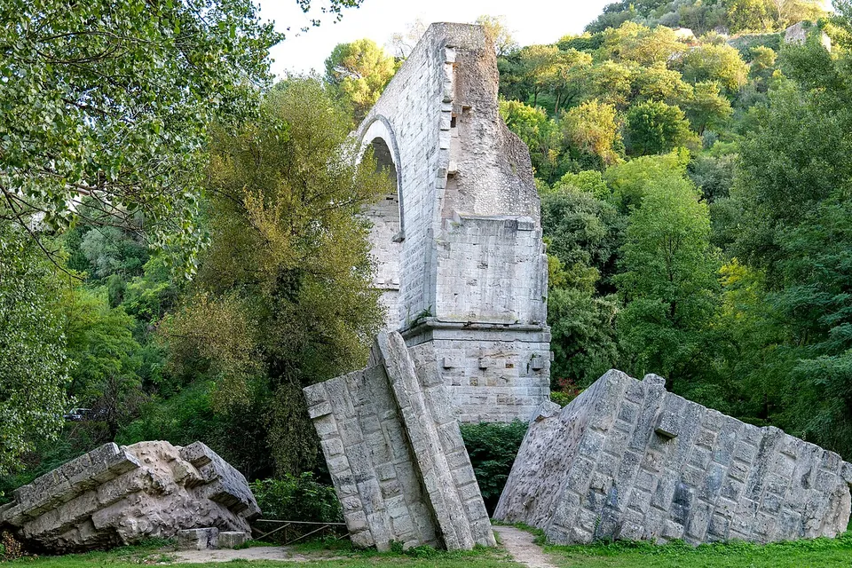 A tall standing stretch of masonry with the start of an arch on its left side, and in front of it great slabs of fallen masonry lying tilted in the grass.