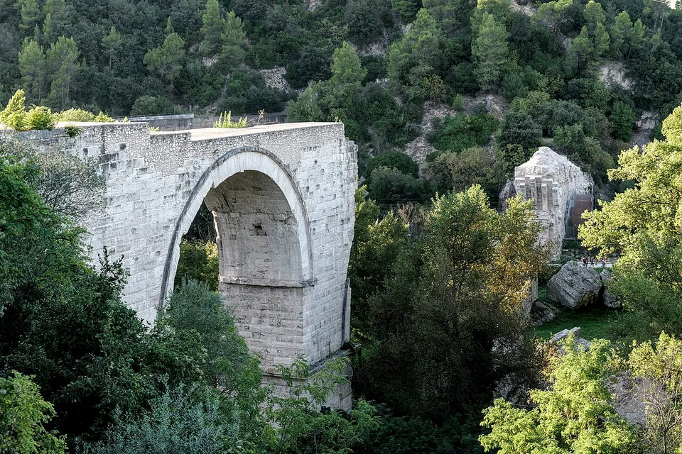 The surviving arch seen from higher ground, rising out of dense trees, with the broken top of another block of masonry standing among the woods to the right.