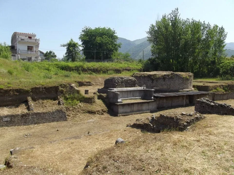 Two large square funerary monuments of grey stone with pale cornices and a flat shelter roof, standing in a dry, grassy excavated area with low walls in front and houses and hills behind.