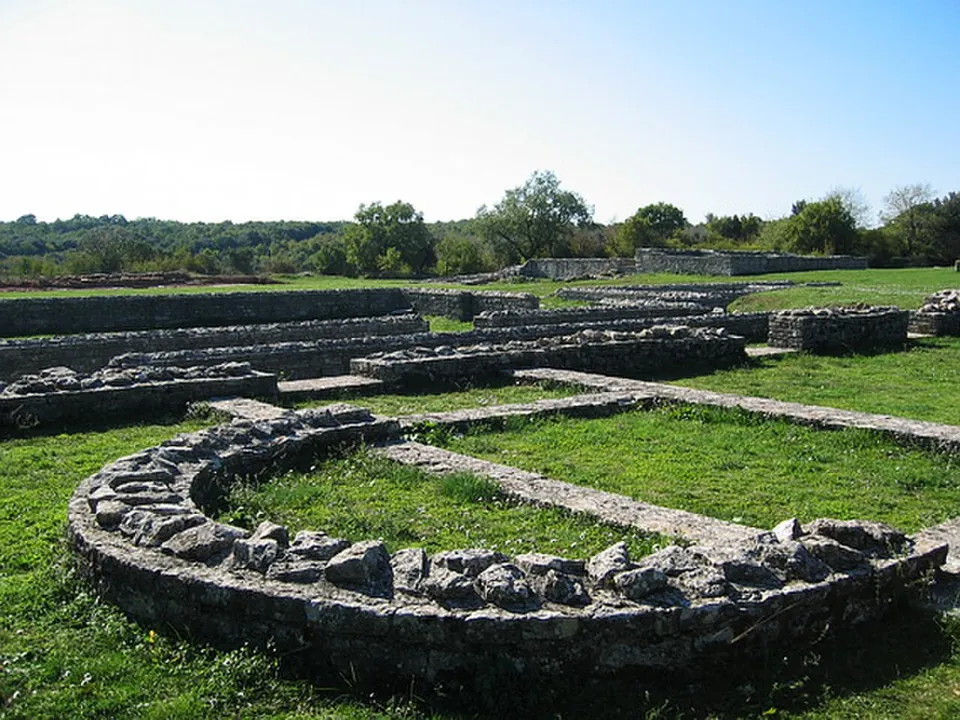 The curved low stone wall of a church apse in the grass in the foreground, with the long parallel wall lines of the nave running away behind it and trees on the horizon.