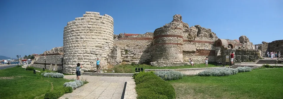 A panorama of ruined fortress walls of stone with bands of brick, a tall round tower on the left and a broken gate tower in the middle, with a paved path, lawns and visitors in front.
