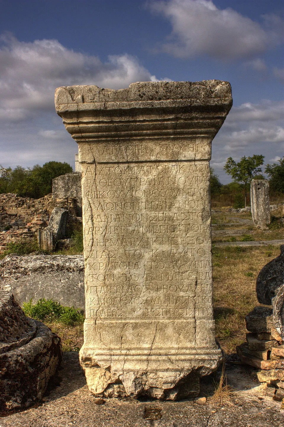 A tall, weathered stone pedestal with a moulded top and base, its face cut with about fifteen lines of Greek capital letters, standing among ruins under a cloudy sky.
