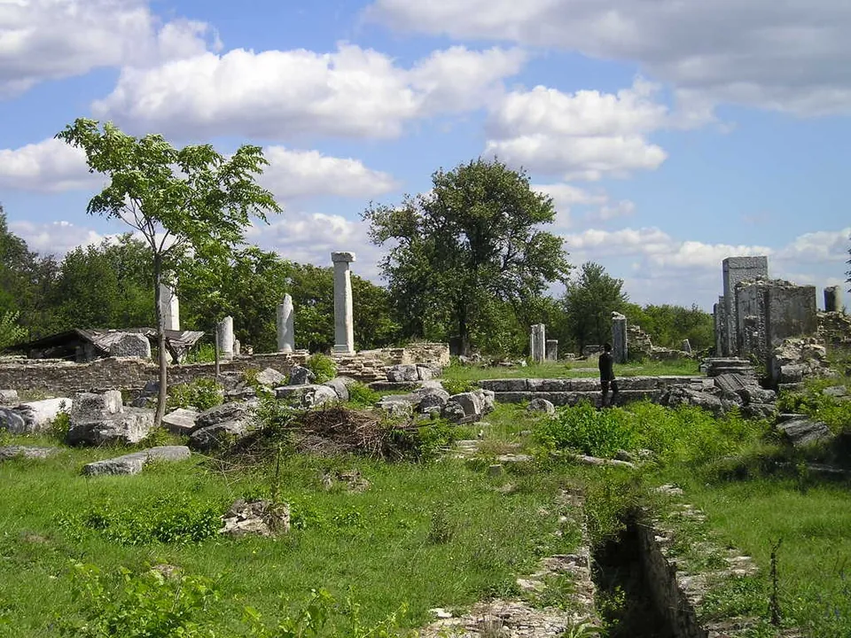 A broad excavated area of low stone walls and fallen blocks in long green grass, with several standing white columns and a tall fragment of wall on the right, trees behind, under a blue sky with white clouds and a visitor walking among the ruins.