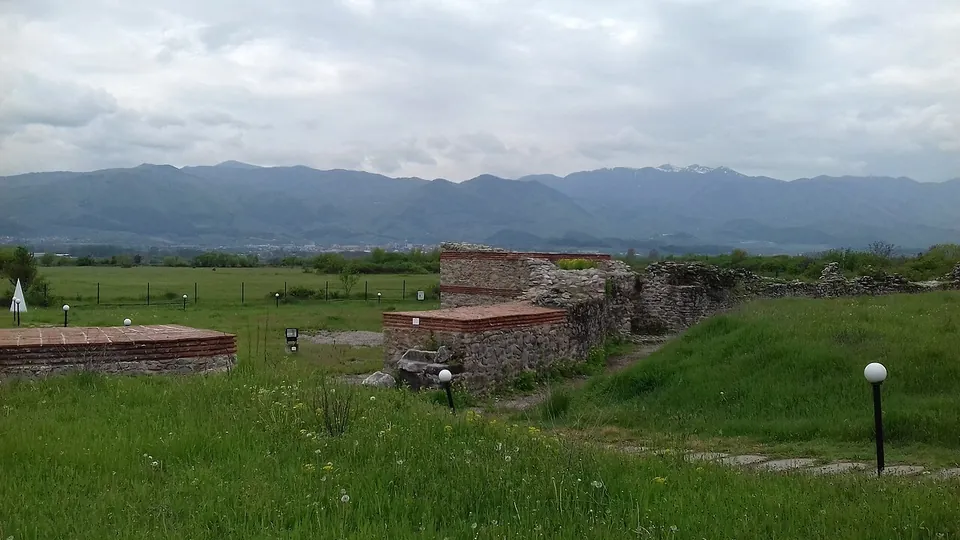 Restored stretches of wall with brick bands and a low brick-capped structure on a grassy site, with lamp posts along a path and a mountain range under clouds behind.